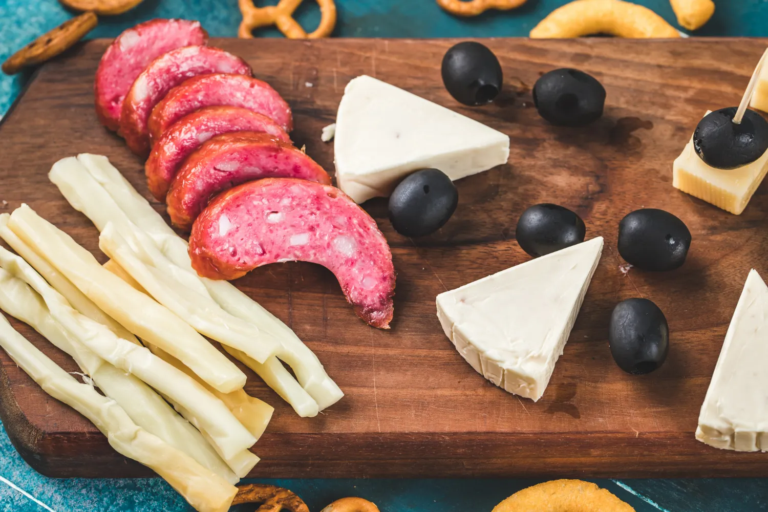 Tabla de embutidos con salchichón, tiras de queso, quesos en forma de triángulo y aceitunas negras, acompañada de aperitivos. Ideal para picnics o reuniones.